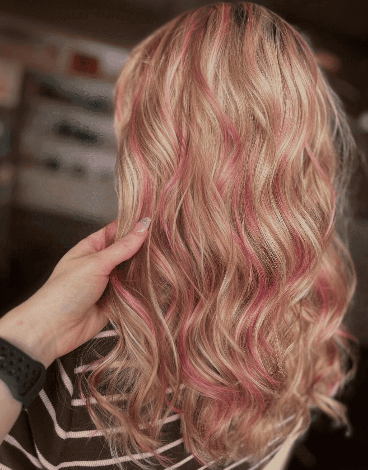 Blonde hair with pink highlights styled in loose waves, held by a hand.