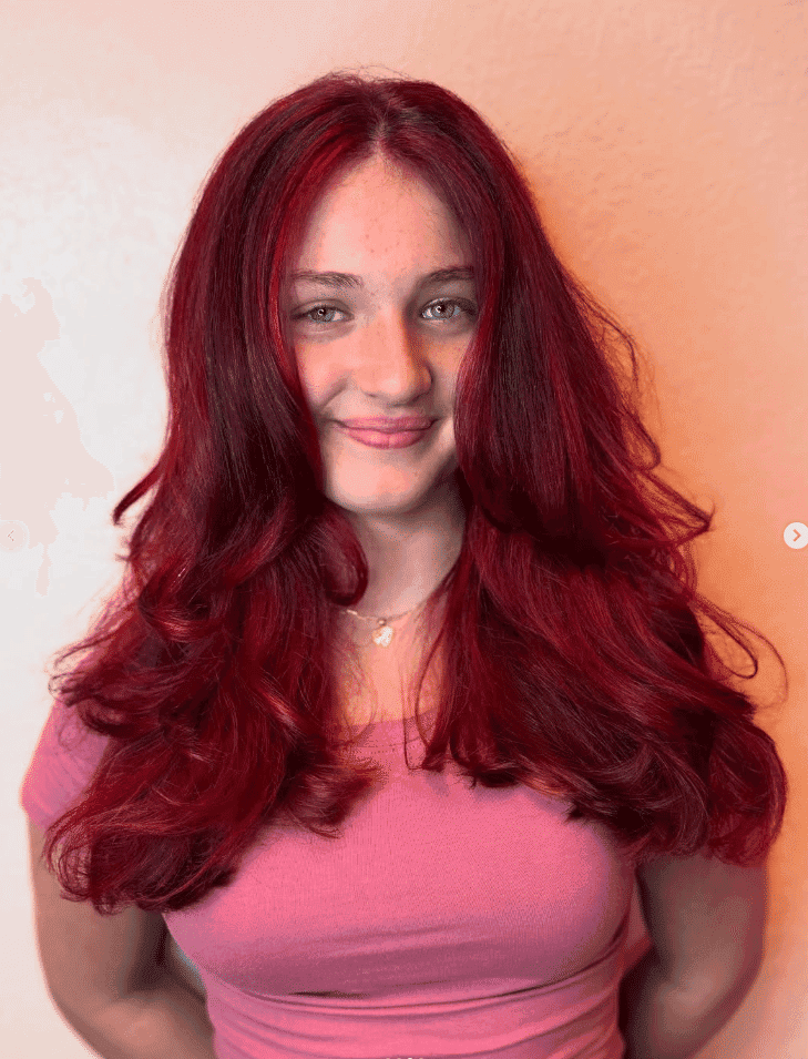 Young woman with vibrant red hair wearing a pink top, smiling against a neutral background.