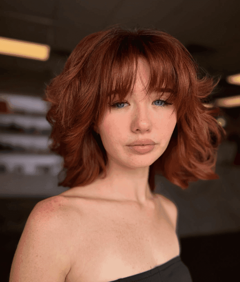 Young woman with red hair and bangs, wearing a strapless top indoors.